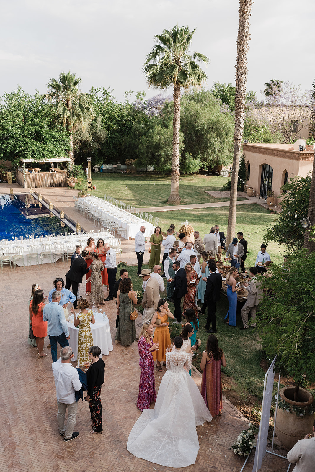 Wedding guests gather outdoors beside a pool and palm trees.
