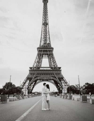 A couple kisses on a wide Paris street with the Eiffel Tower behind them, photographed in black and white.