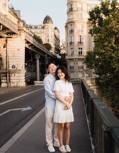 One partner embraces the other from behind on a walkway beside Bir-Hakeim Bridge.