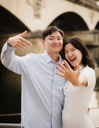 The couple smiles at the camera and holds a hand forward to show the engagement ring.