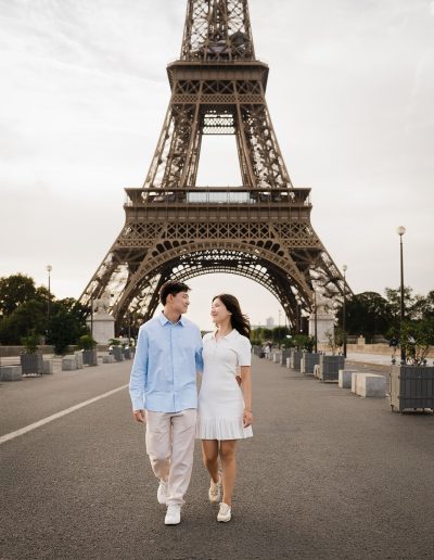 A couple walks together on a quiet road with the Eiffel Tower rising behind them.