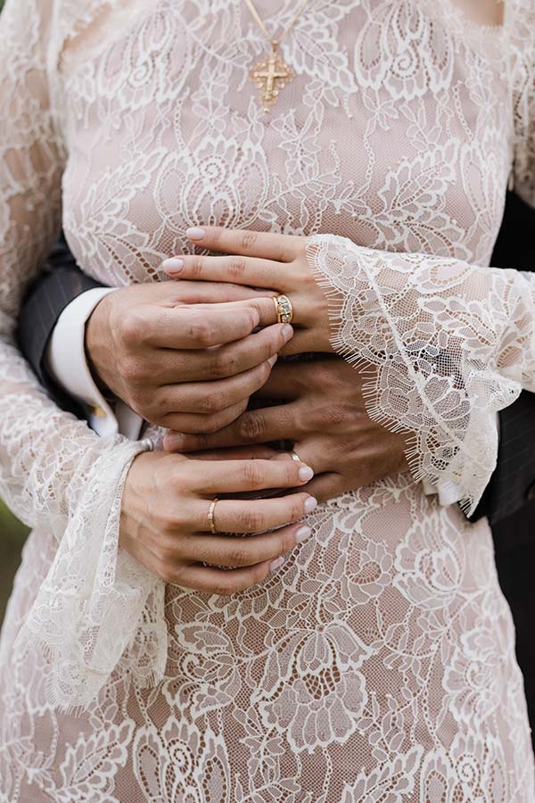 Gros plan sur les mains et les alliances d’un couple posées sur une tenue en dentelle blanche.
