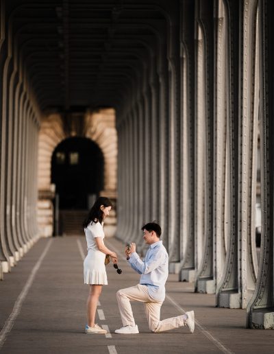 A person proposes on one knee beneath Bir-Hakeim Bridge while their partner stands facing them.