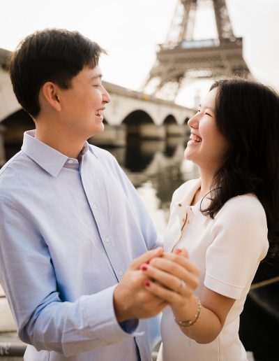 A newly engaged couple smiles at each other while holding hands near the Eiffel Tower.
