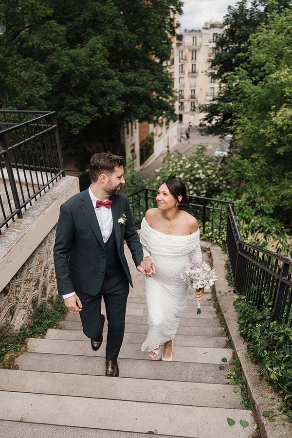 Un couple en tenue de mariage monte un escalier extérieur à Montmartre main dans la main, bouquet à la main.