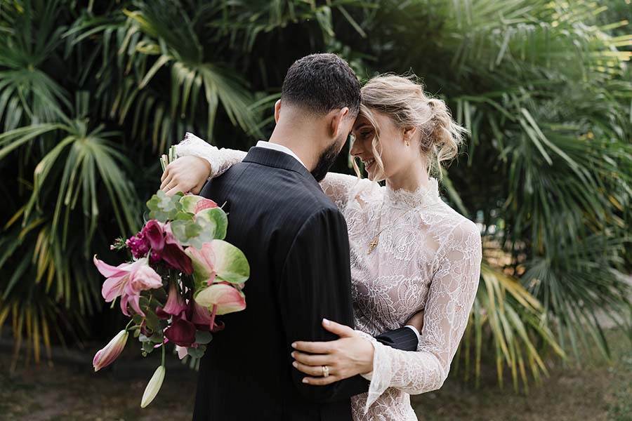 Un couple s’enlace devant des feuillages, avec un bouquet de fleurs roses et pourpres.