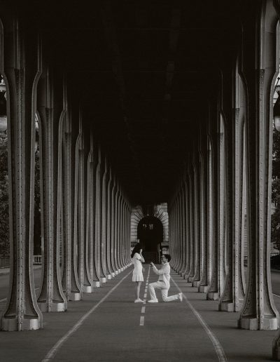 A person kneels to propose beneath the arches of Bir-Hakeim Bridge in Paris, photographed from a distance in black and white.