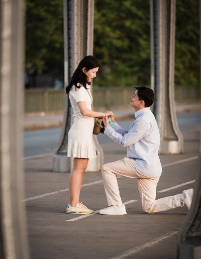 Close view of a person proposing on one knee and holding their partner’s hands beneath the bridge.